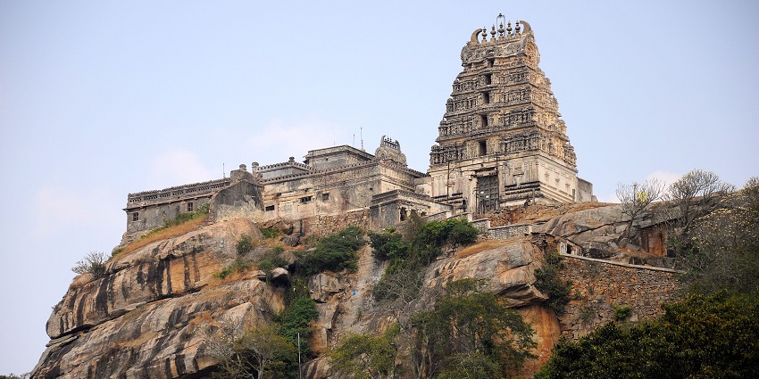 An ancient stone temple complex atop a rocky hill, with green surroundings and skies.
