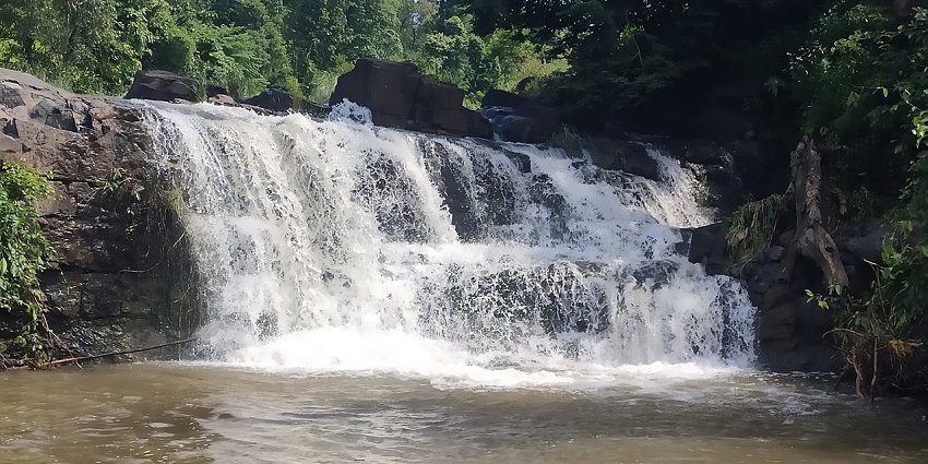 A small waterfall cascading over dark rocks into a calm, shallow pool surrounded by greenery