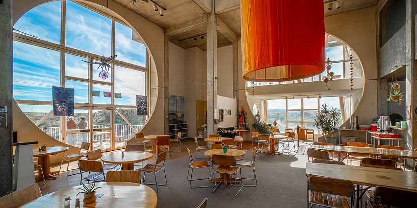 Modern café with tall concrete walls, round windows, and wooden tables under an orange drape, one of the best cafes in Kalimpong.
