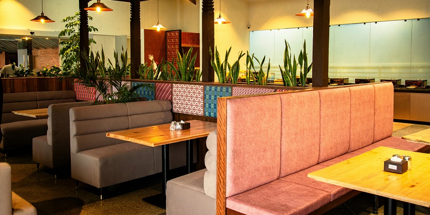 Restaurant interior with cushioned booths, wooden tables, hanging lamps, and plants.
