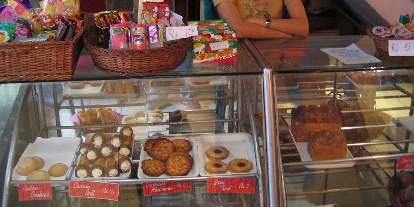 Cosy café interior with pastry display, hanging lights, and shelves stocked with drinks.

