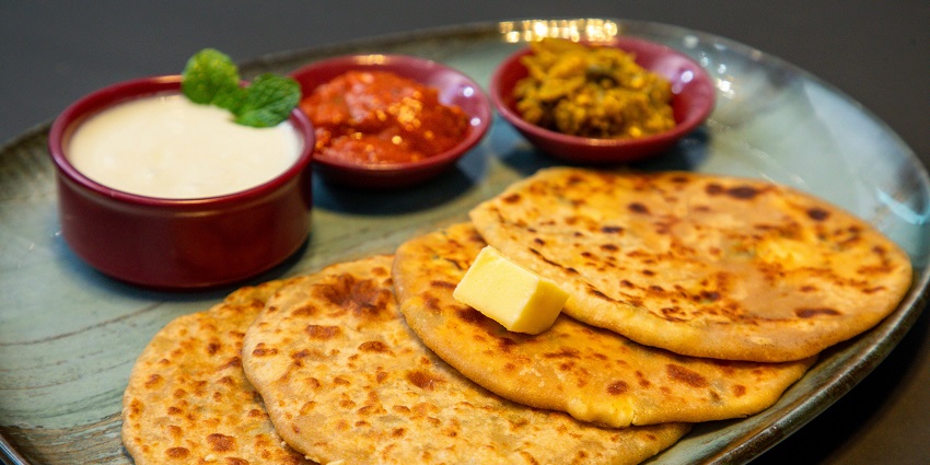 A plate of aloo paratha is served with a bowl of curd and achaar on a wooden table.