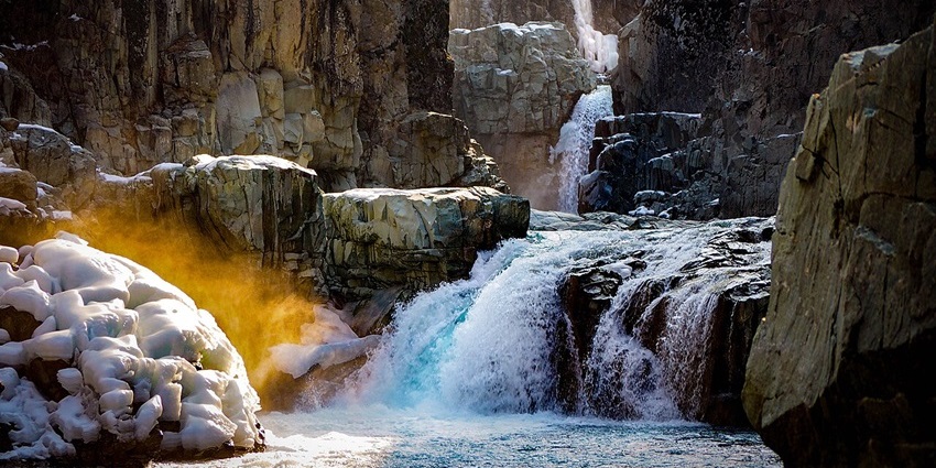 A sunlit waterfall cascades over a rocky cliff face into a pool, surrounded by snow and ice.