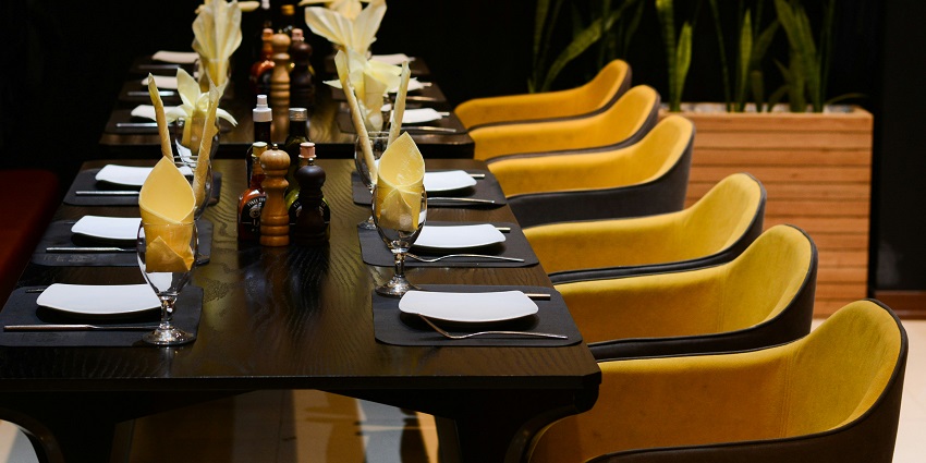 Ambient Cafe interior featuring a long table with yellow chairs and black table cloths.

