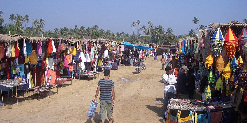 A busy, outdoor market with many stalls selling clothing and goods, in a dusty field.