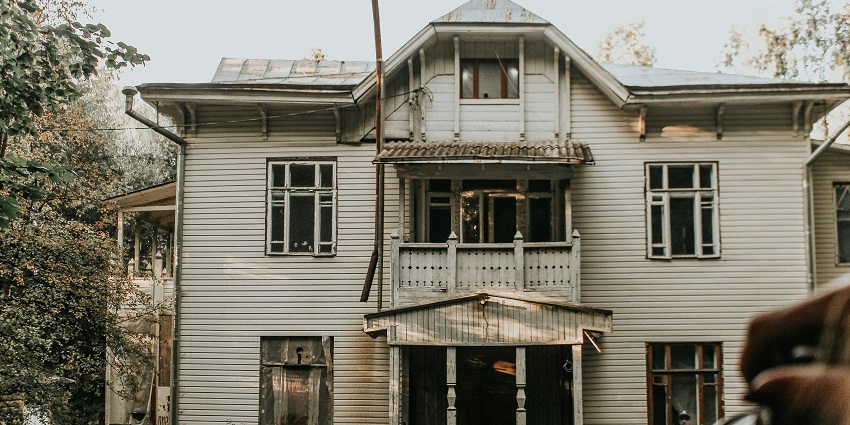 Old abandoned house with weathered walls surrounded by dry grass and trees.