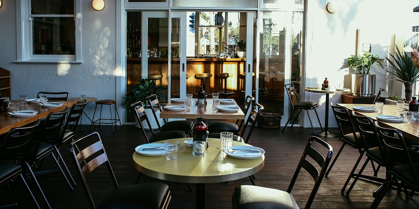 Bright restaurant dining room with set tables, black chairs, and sunlight streaming inside.