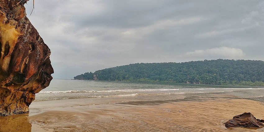 Beach of Bako National Park with a big rocky structure and low waves hitting the sandy shores.
