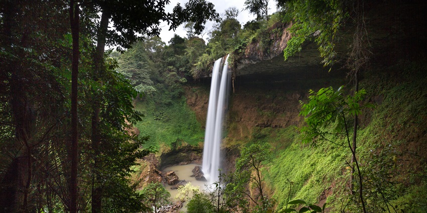 A tall waterfall plunges from a mountain cliff surrounded by mist, rocks, and greenery.