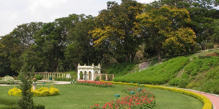 A lush green park with a white building and curved flowerbeds sits under a partly cloudy sky.