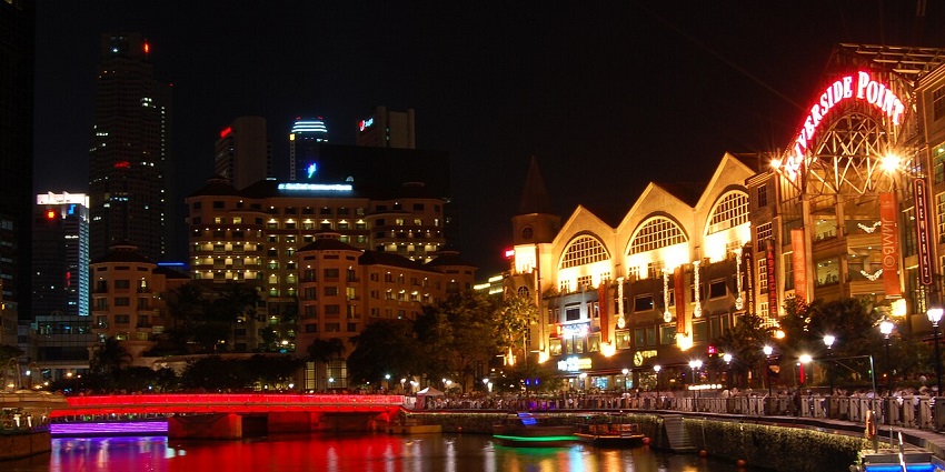 People dining outdoors by the riverside at night with city lights in the background

