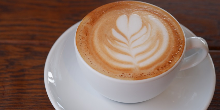 A steaming cup of latte with creamy foam, leaf design in the foam on a wooden table.