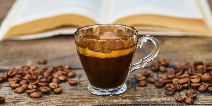 Glass cup of espresso with coffee beans on a wooden table and an open book in the background
