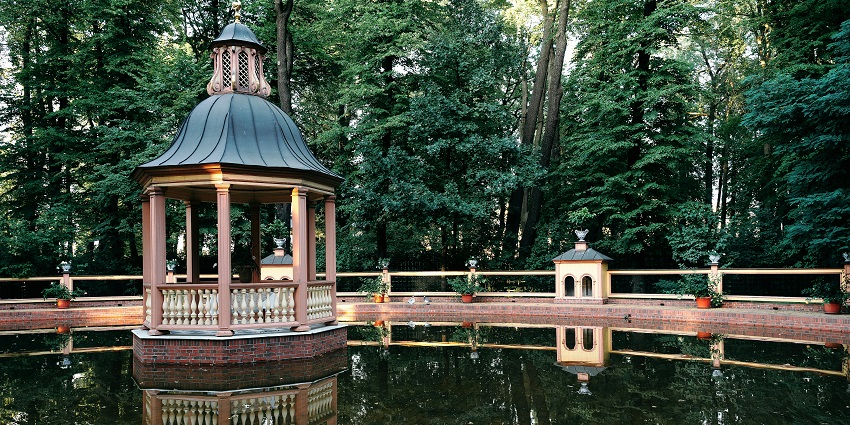 Reflections of a hut on calm water surrounded by tall trees at Campal Gardens, one of the top parks in Goa.
