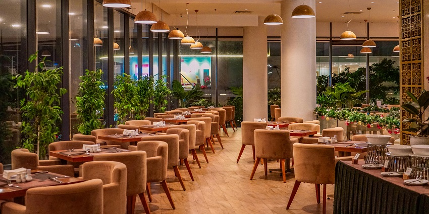 Restaurant with plush beige chairs, hanging dome lights, and greenery along tall windows.