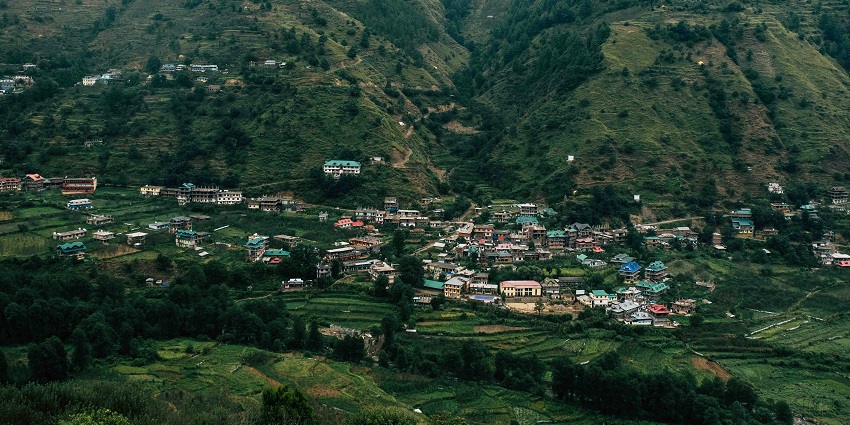 View over Chamba, Himachal Pradesh, showing green slopes, river, and mountains.
