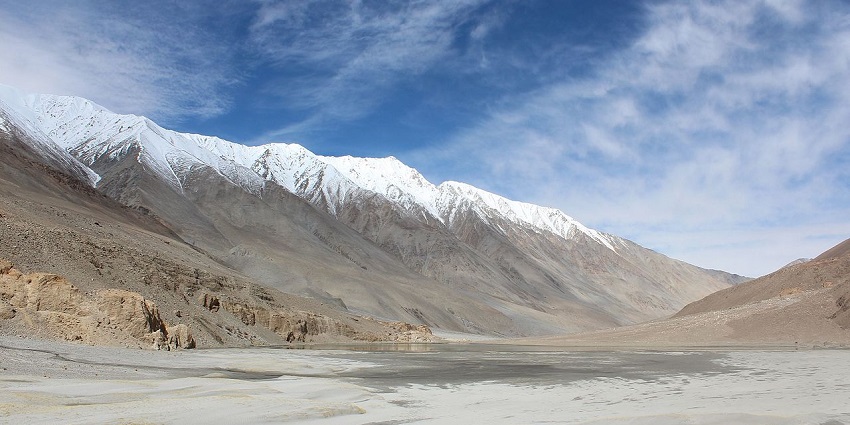 A wide valley with a small stream leading to snow-capped mountains under a blue sky.
