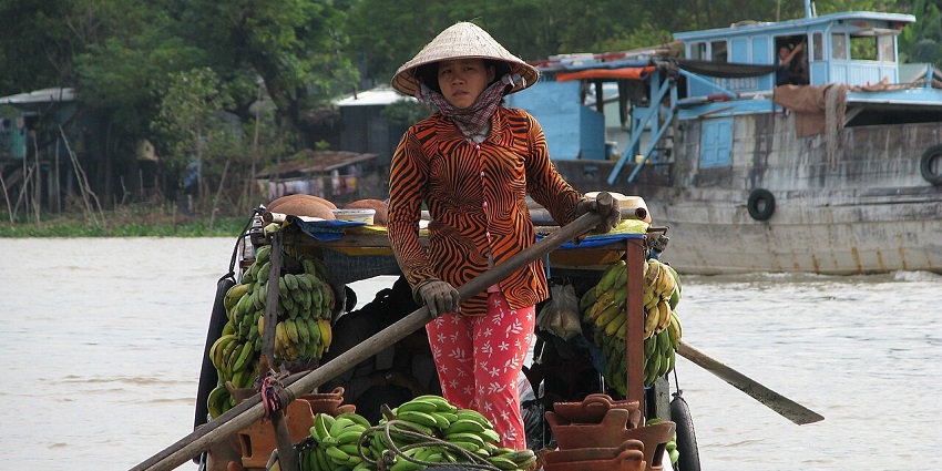 A woman in a conical hat stands in a boat filled with bananas and clay pots, sailing.
