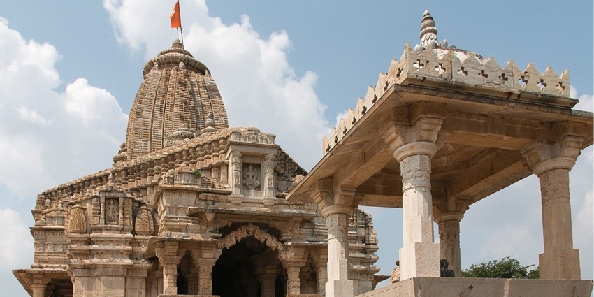 A stone temple with an intricate, multi-tiered roof sits on a hill, with green trees in the foreground.