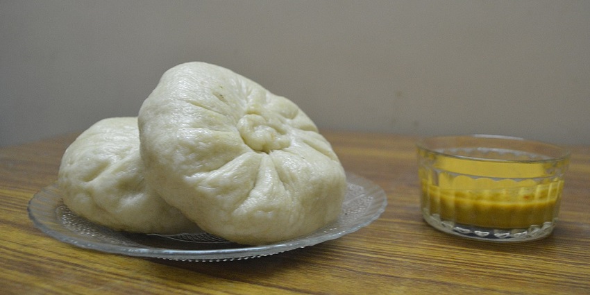 A large, round steamed dumpling with pleated folds at the top, set on a glass plate.
