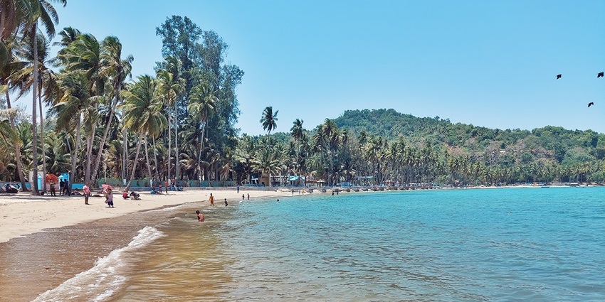 A scenic view of Corbyn’s Cove Beach with people walking on the sandy beach with palm trees and ocean.