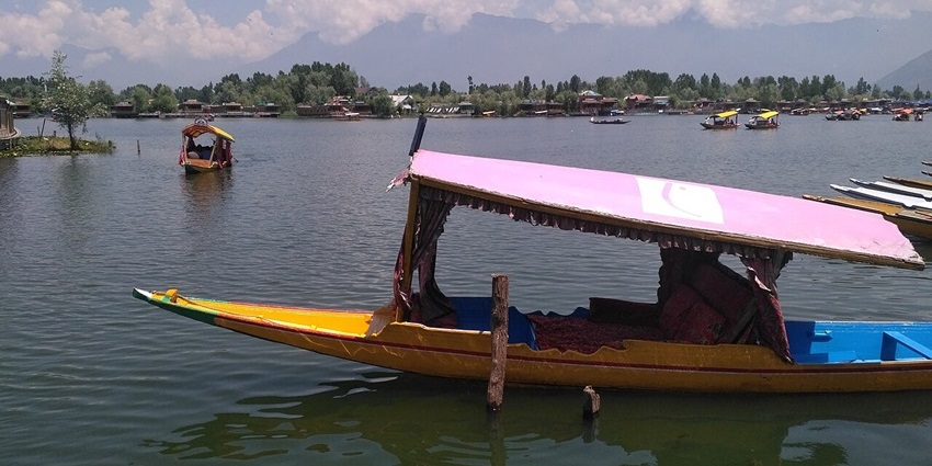 A long, yellow boat with a pink canopy floats on a calm lake, with mountains in the distance.