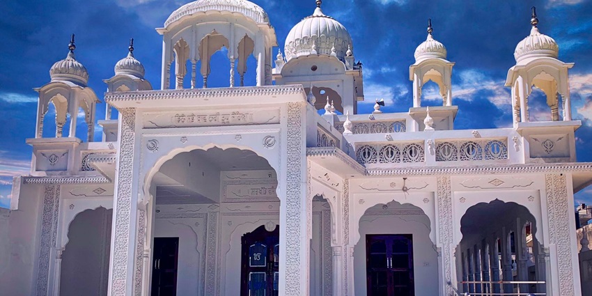 A white marble gurdwara with ornate domes and arches stands against a dramatic blue sky with clouds.