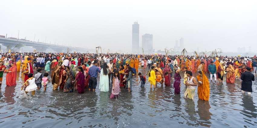 People standing in the Yamuna River celebrating Chhath Puja next to a bridge and high-rise buildings behind.
