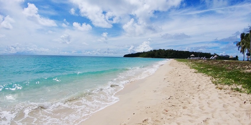 A wide, white sandy beach with light blue water and gentle waves under a cloudy sky.