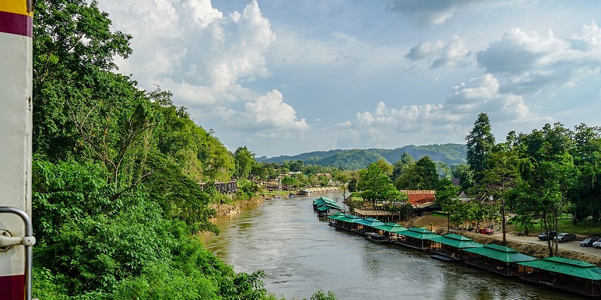 River flowing through lush hills and riverside huts at Erawan National Park, Thailand.
