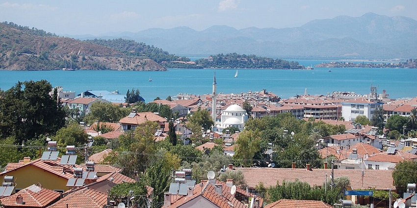 An aerial view of a coastal town with red-roofed houses, a mosque, and a calm bay.
