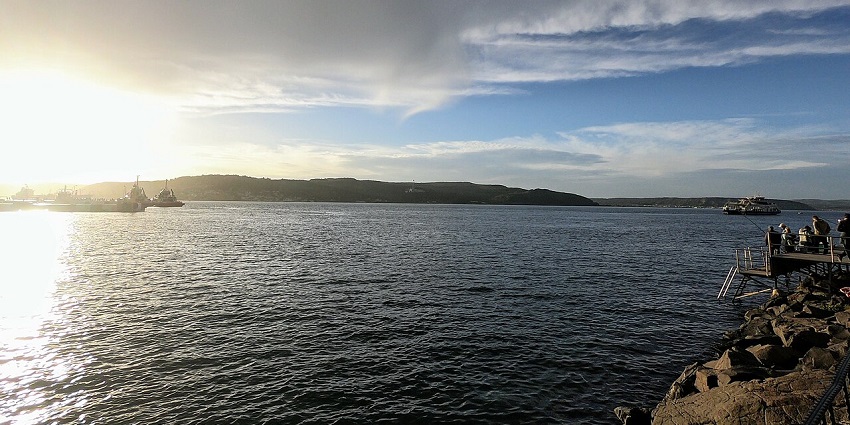 A moody, wide-angle shot of a harbour at sunset, with boats and a rocky shoreline.
