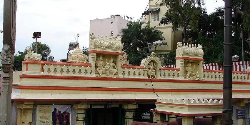 A temple with a yellow and orange facade, adorned with statues and decorative roof carvings, under a cloudy sky.
