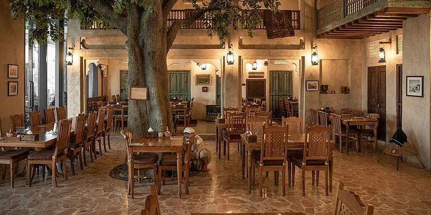 A rustic restaurant with many wooden tables and chairs, and a large tree in the centre.

