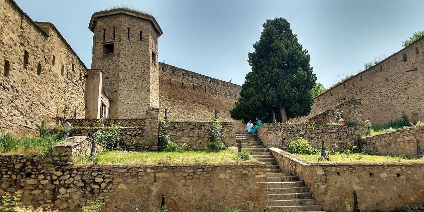 A wide, panoramic view of a fort with several distinct sections on a hilltop, surrounded by a city, one of the best forts in Kashmir.