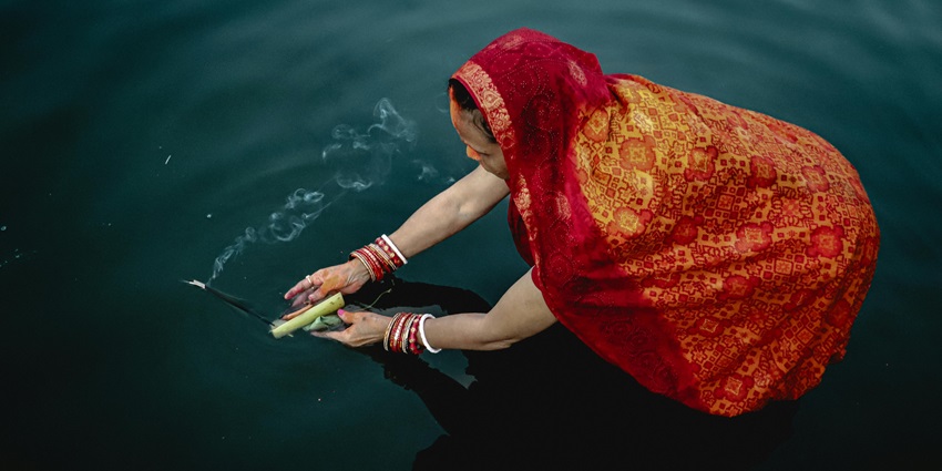 A woman in a saree holding incense and leaves while partially soaking in the water during Chhath Puja.