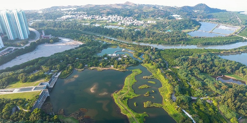 An aerial view of a winding body of water with small islands surrounded by green fields, buildings, and hills, one of the zoos in Hong Kong.