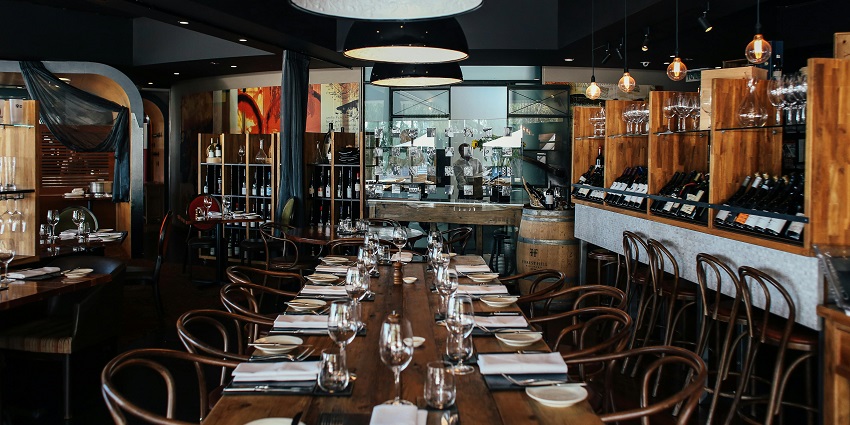 Long wooden dining table set with glassware, wine shelves, and hanging black lamps above
