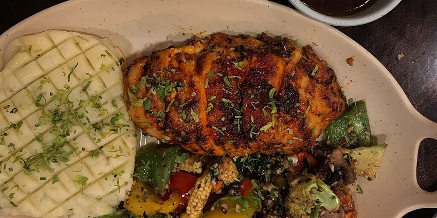 A platter of food with grilled chicken, mashed potatoes, mixed vegetables, and a side of sauce.
