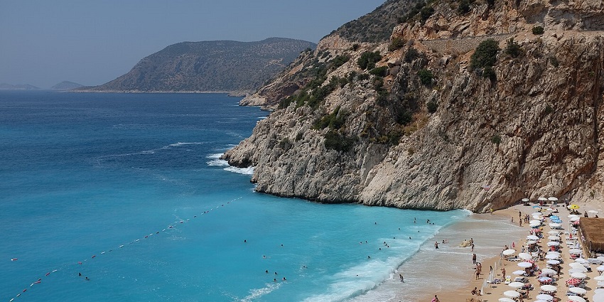 A vibrant blue beach with umbrellas and swimmers below a cliff, perfect for scuba diving in Turkey.
