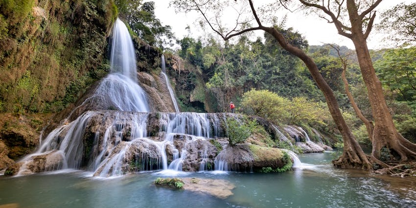 Powerful waterfall rushing through dense green forest, creating mist across the valley.
