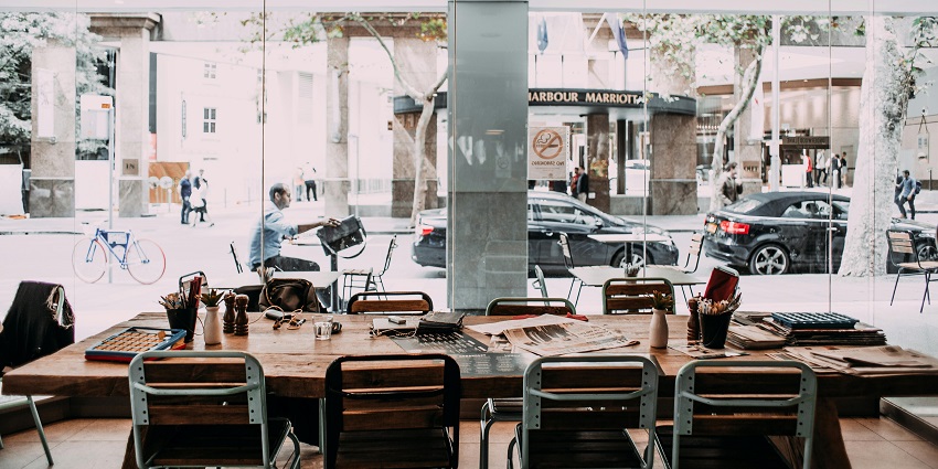 Café with a long wooden table, metal chairs, newspapers, and large glass windows facing the street.
