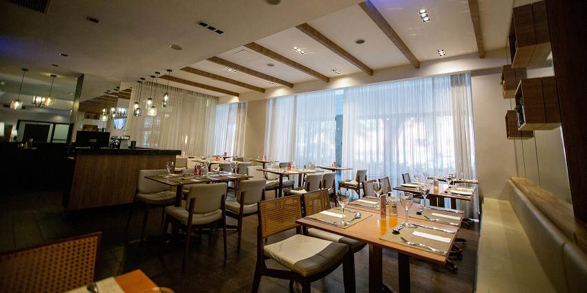 Bright restaurant with wooden tables, cushioned chairs, and sheer curtains along windows
