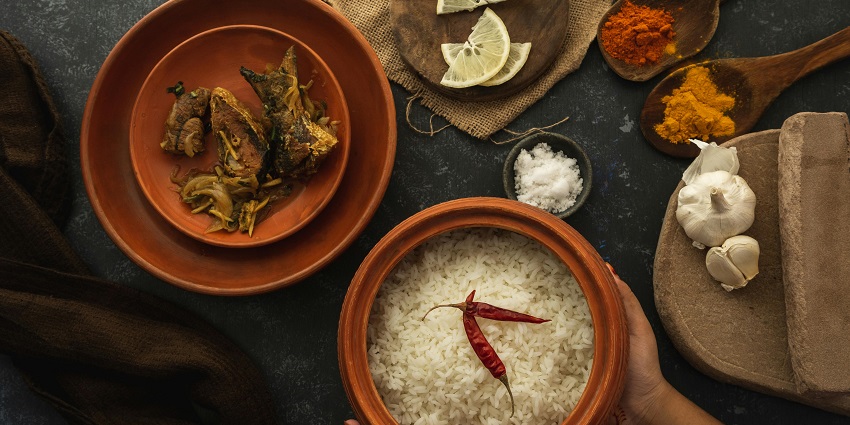 A flat-lay view of a meal with a clay bowl of white rice, a plate of fish, and a few spices and lemons.
