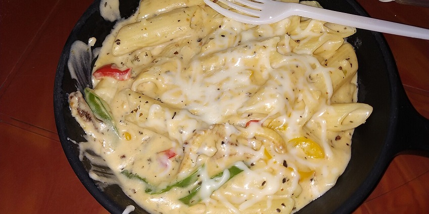 Hot skillet of creamy cheese pasta with peppers, served with a white fork and seasoning in one of the restaurants in City Centre Siliguri.
