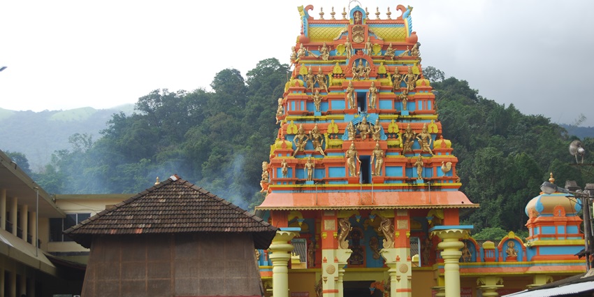A vibrant, colourful temple with a tiered tower stands next to a wooden building under a grey sky.