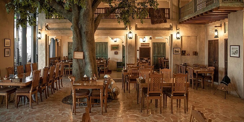 A rustic restaurant with many wooden tables and chairs, and a large tree in the centre.
