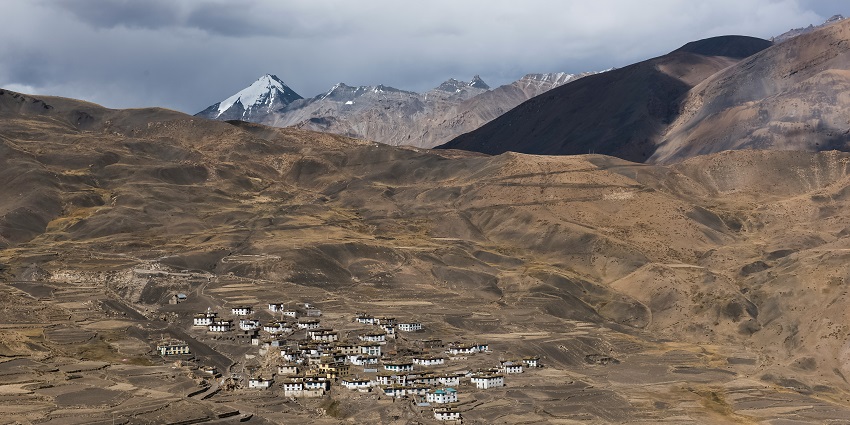 View of Thenu village in Lahaul showing farmland, riverbank, and mountains in distance.
