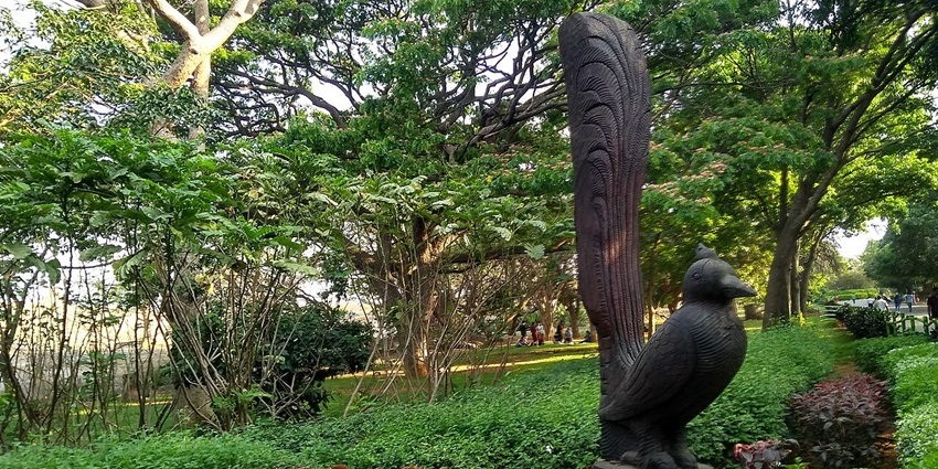 A carved stone peacock statue stands in a garden with dense foliage and large trees.