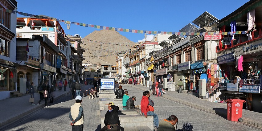 Bustling Leh market street with prayer flags, shops, and a huge mountain backdrop.
